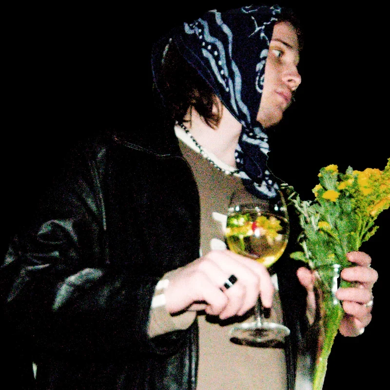 Kéquetz wearing a patterned headscarf while holding yellow flowers and a glass